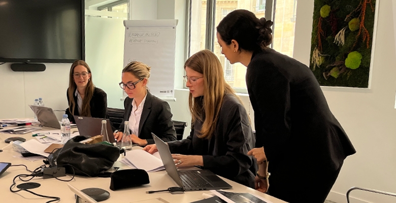 Women in professional attire working together around a table with laptops, documents, and water bottles, engaged in a collaborative consulting session in a modern office environment.