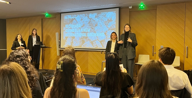 Women presenting in a conference room with a slide titled "Recycling: Giving Life a New International Beginning," while an audience listens attentively.