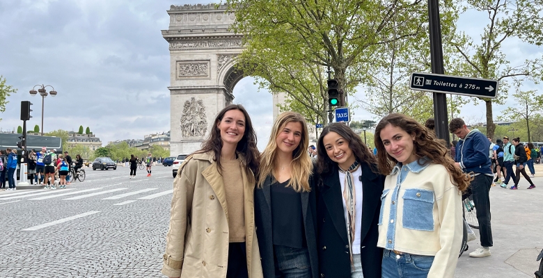 Four young women standing smiling near the Arc de Triomphe in Paris, dressed in casual stylish clothes, with trees and city street in the background after enjoying the Own Your Future experience.