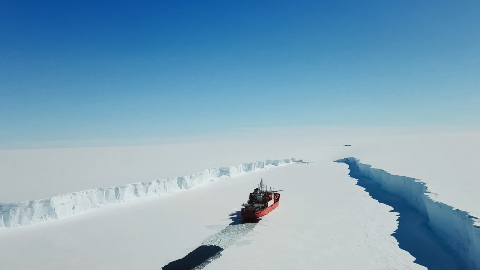 A ship breaking through a vast ice sheet in a polar region under a clear blue sky.