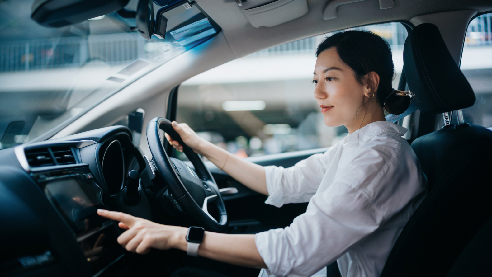 A person wearing a white shirt and smartwatch is sitting in the driver's seat of a car, reaching to touch the dashboard screen.