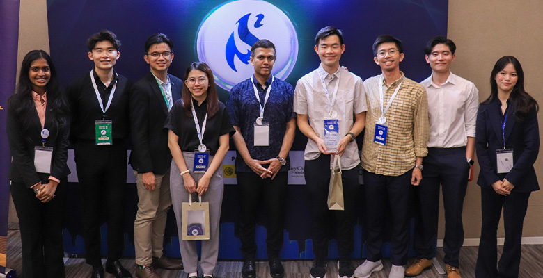 Nine people in formal attire stand with certificates and gift bags at an award event, posing in front of a branded backdrop with conference badges.