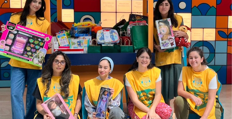 A group of children, alongside adults, is standing in a brightly colored area holding toys and games, participating in a charity event to distribute gifts.