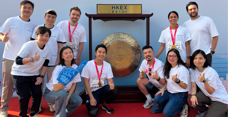 A group of people is shown posing together in matching white t-shirts after completing a charity run event, celebrating their participation with medals in front of a large gong.