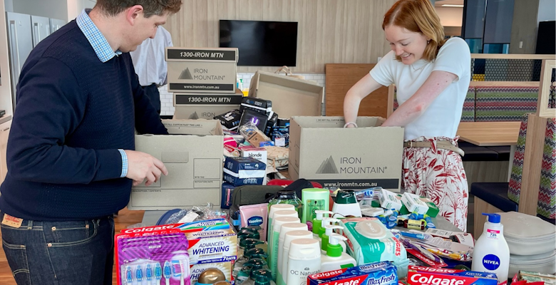 Two individuals are sorting through boxes filled with personal care items, such as toothbrushes and soaps, in an office environment, contributing to a community aid initiative.