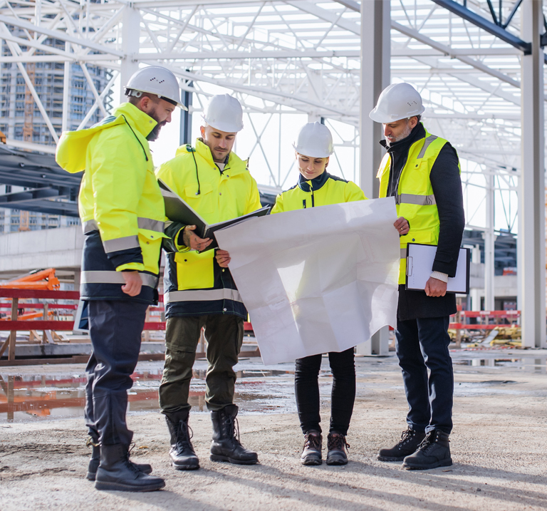 A group of four construction workers in bright yellow vests and helmets review plans on a construction site.