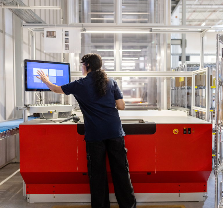 A worker interacts with a touchscreen monitor in a warehouse, overseeing automated processes beside a blue conveyor belt.
