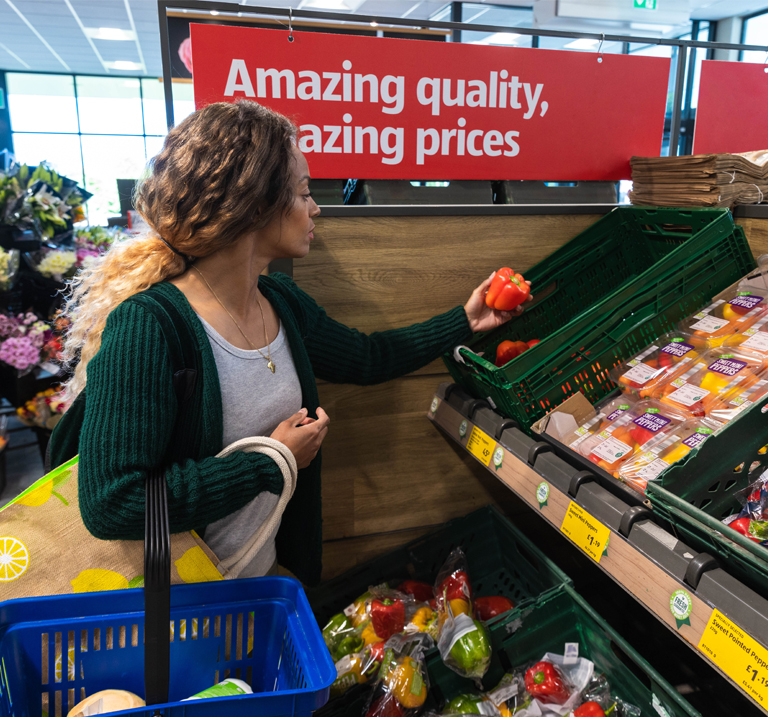 A shopper inspects colorful vegetables at a market, with a sign highlighting amazing quality and prices in the background.