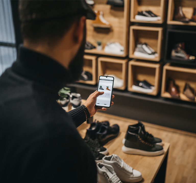 A man with a beard examines shoe options on his smartphone in a stylish shoe store, with various sneakers displayed in the background.