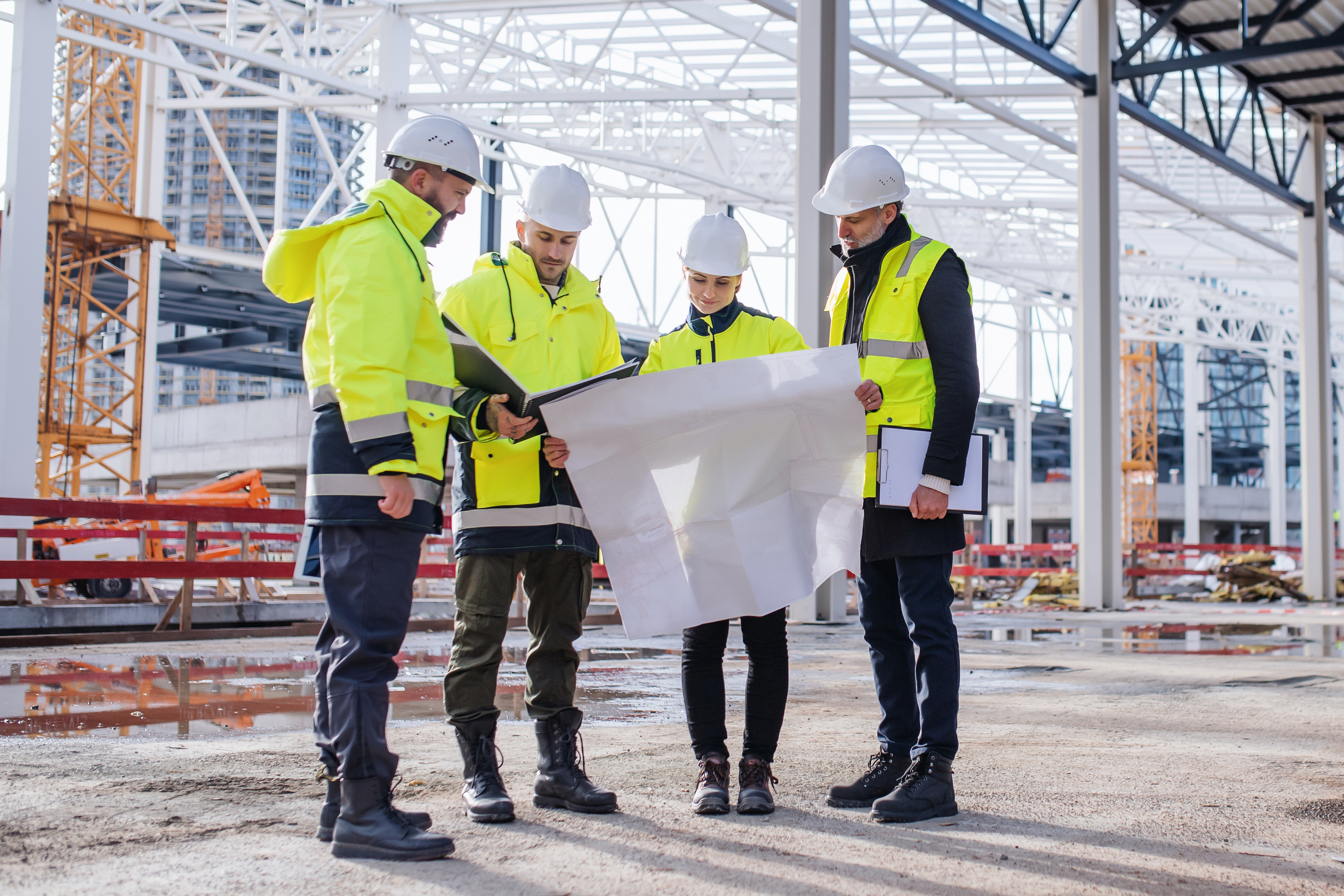 A group of four construction workers in bright yellow vests and helmets review plans on a construction site.