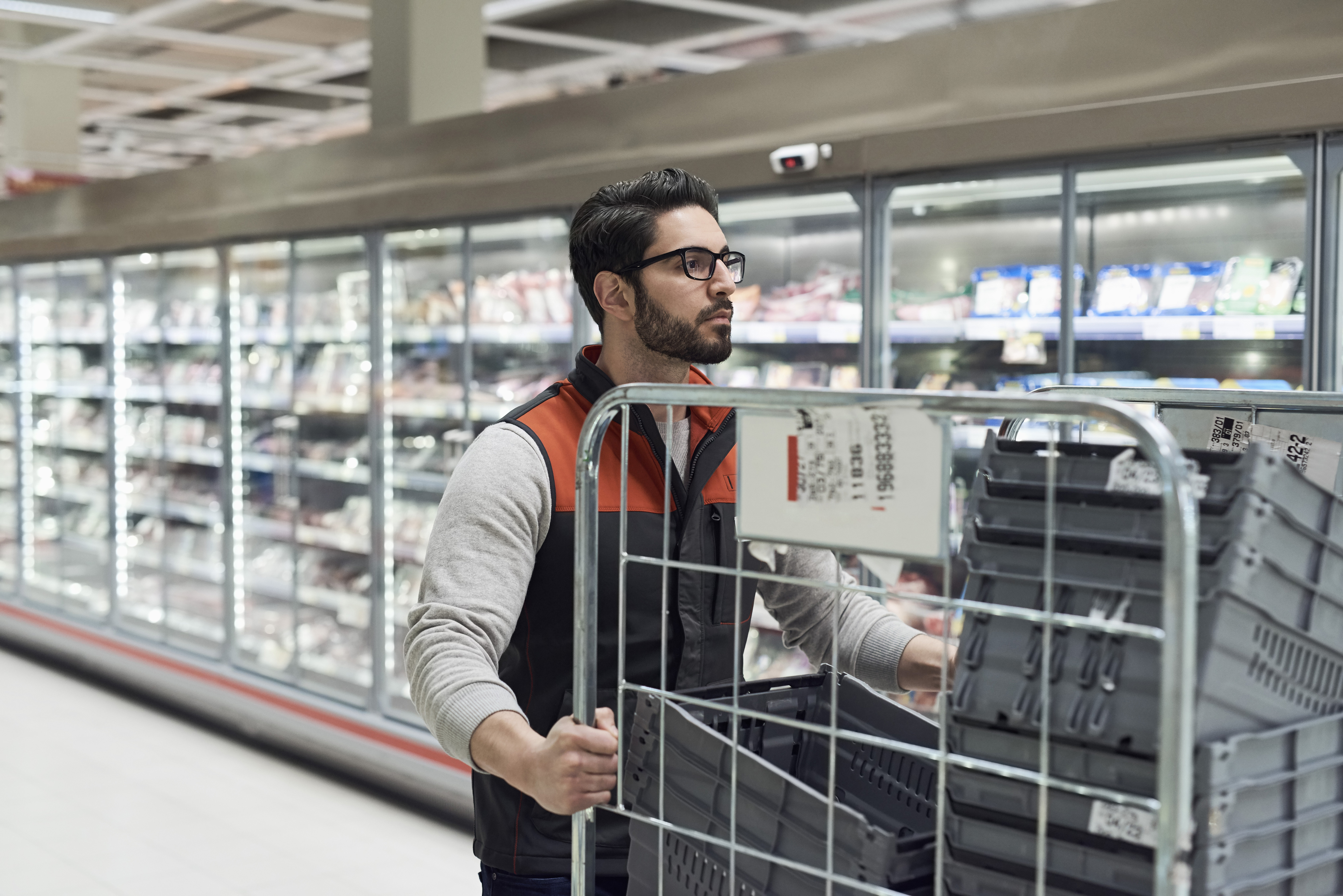 A person in a black and red vest pushes a cart filled with empty shopping baskets in a grocery store aisle with refrigerated displays.