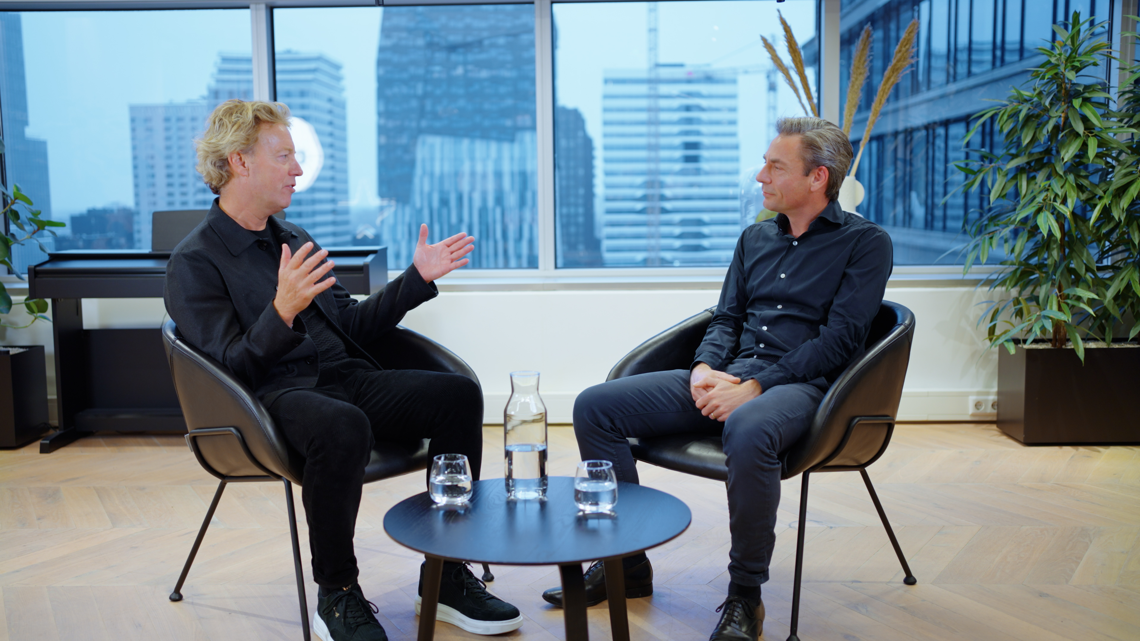 Two professionals sit on a modern couch in an elegant interior, engaged in conversation with soft lighting and green accents.