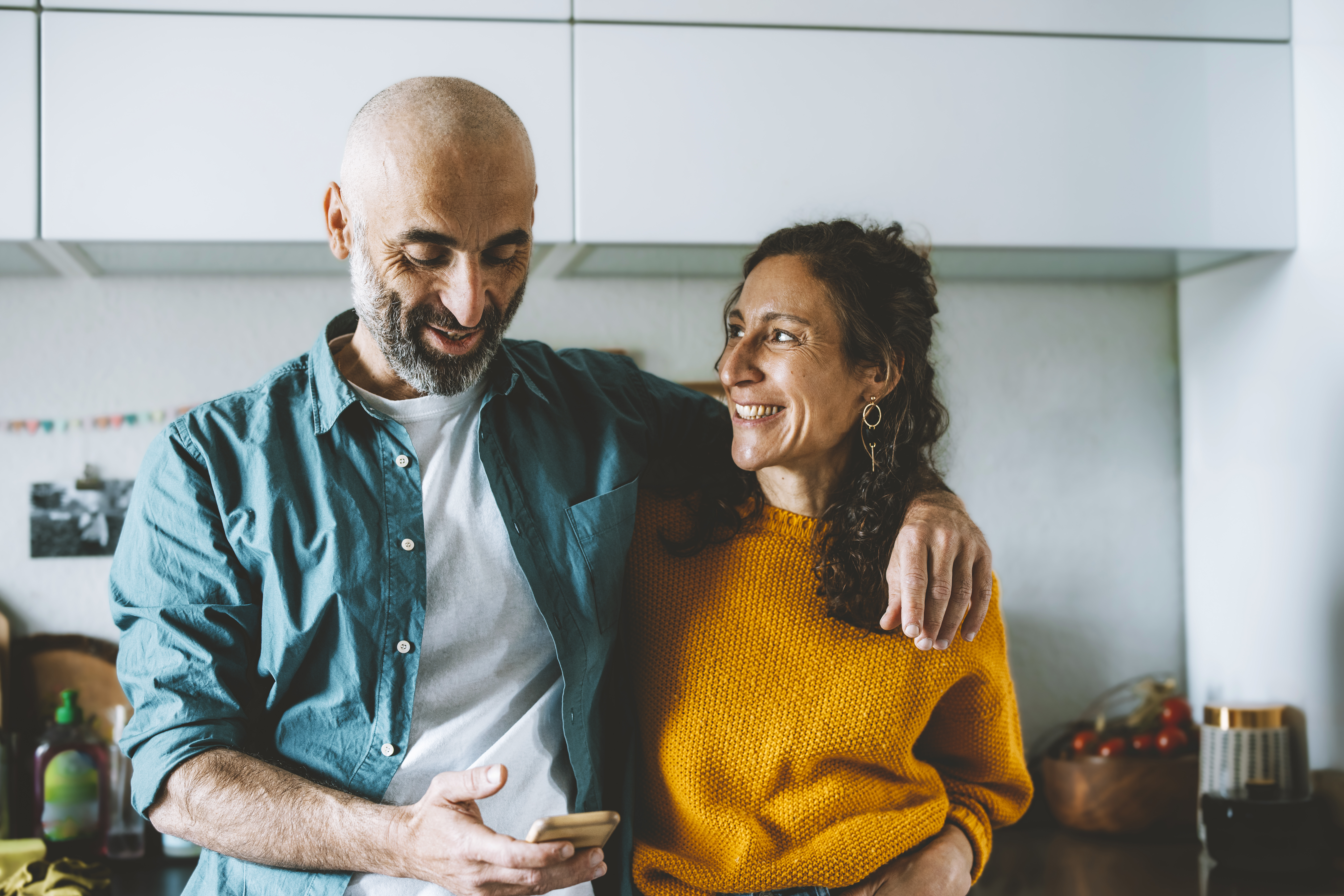 A couple stands close together, smiling, with one holding a phone, in a bright kitchen setting with modern decor.