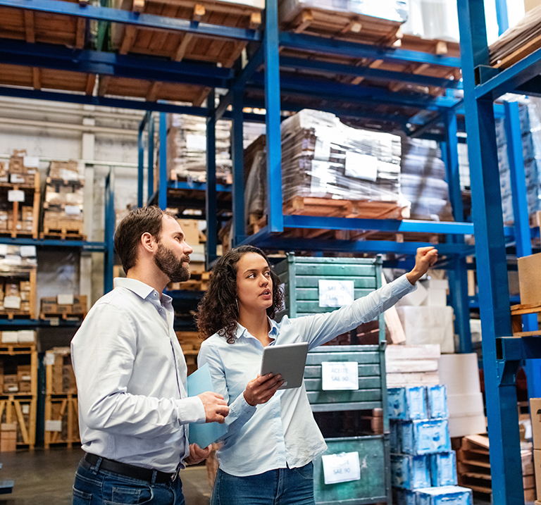 Two people in business attire stand in a warehouse; one holds a tablet while the other points toward shelves stacked with boxes and pallets.