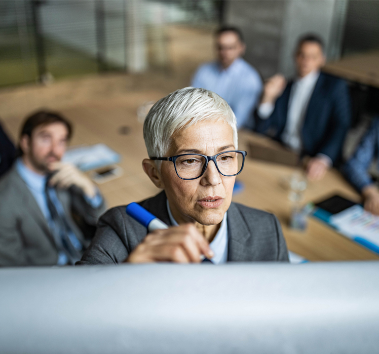 An individual with short gray hair writes on a whiteboard during a meeting, with four people seated at a table in the background.
