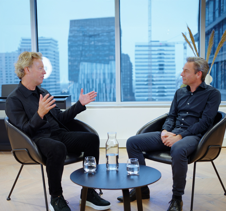 Two professionals sit on a modern couch in an elegant interior, engaged in conversation with soft lighting and green accents.