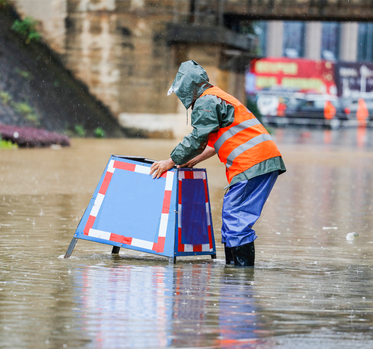 A worker in waterproof gear sets up a warning sign in a flooded street, heavy rain falling around him.
