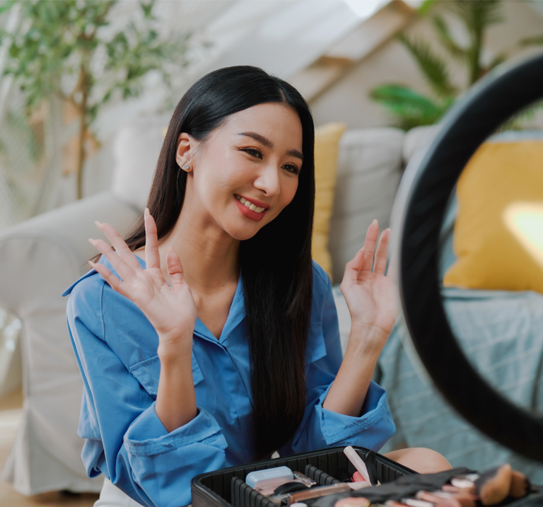 Person in a blue shirt faces a ring light, smiling with open hands. A makeup kit is visible in front. Vibrant living room setting.