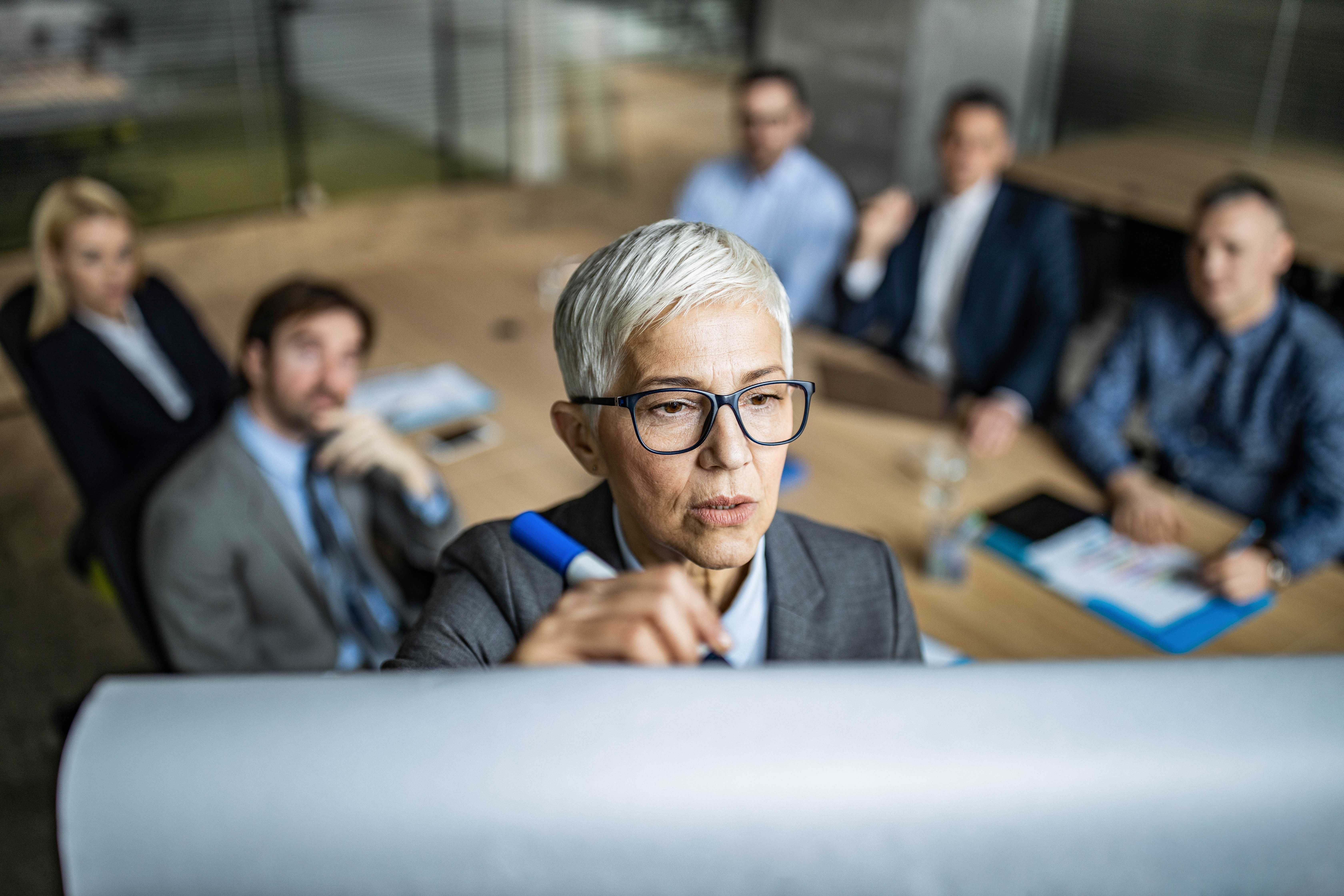 An individual with short gray hair writes on a whiteboard during a meeting, with four people seated at a table in the background.