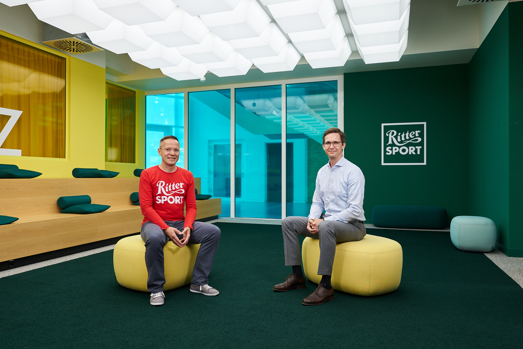 Two men sit on round ottomans in a modern, colorful office space with a Ritter Sport logo on a green wall behind them. One wears a red Ritter Sport shirt; the other wears business attire.