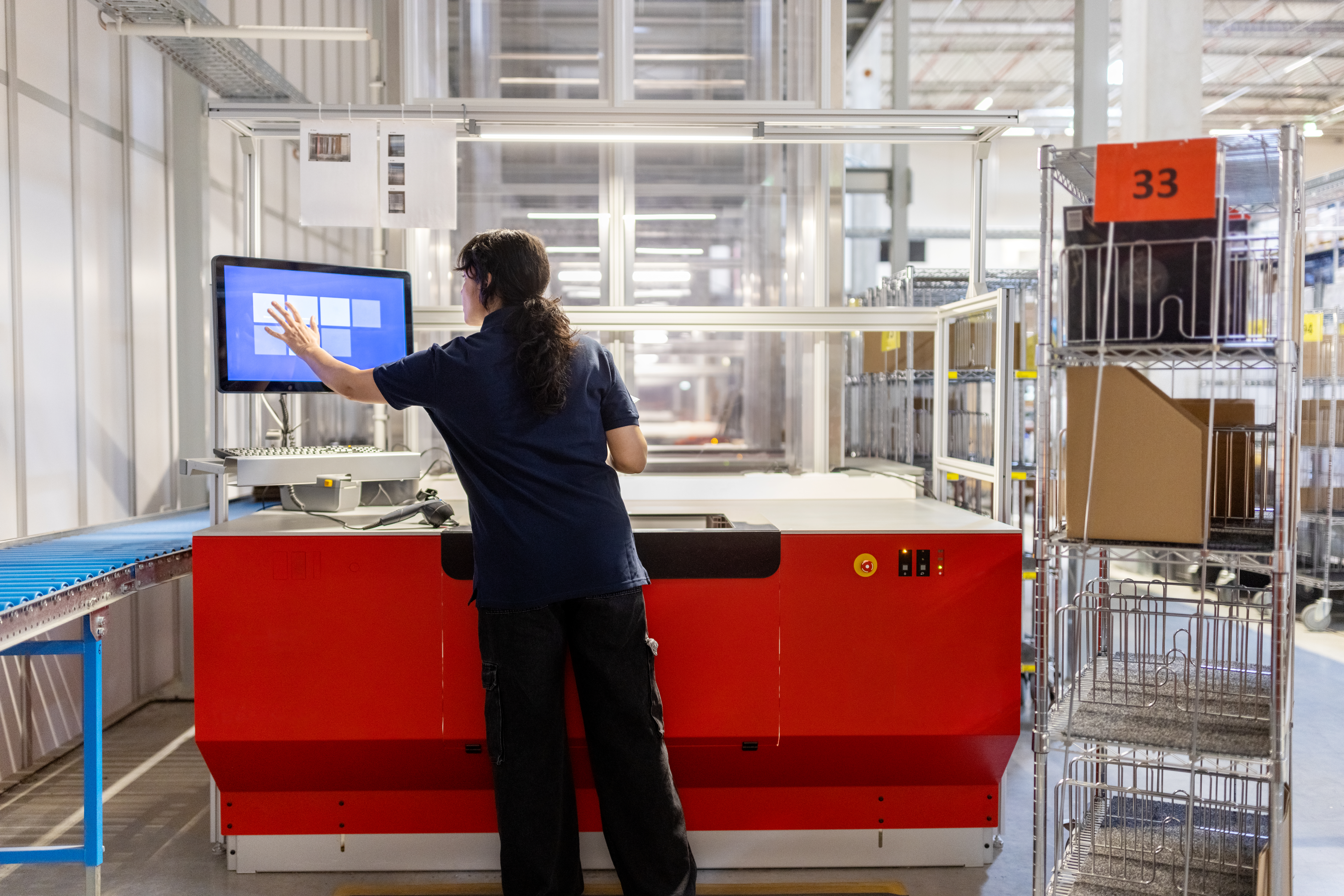 A worker interacts with a touchscreen monitor in a warehouse, overseeing automated processes beside a blue conveyor belt.