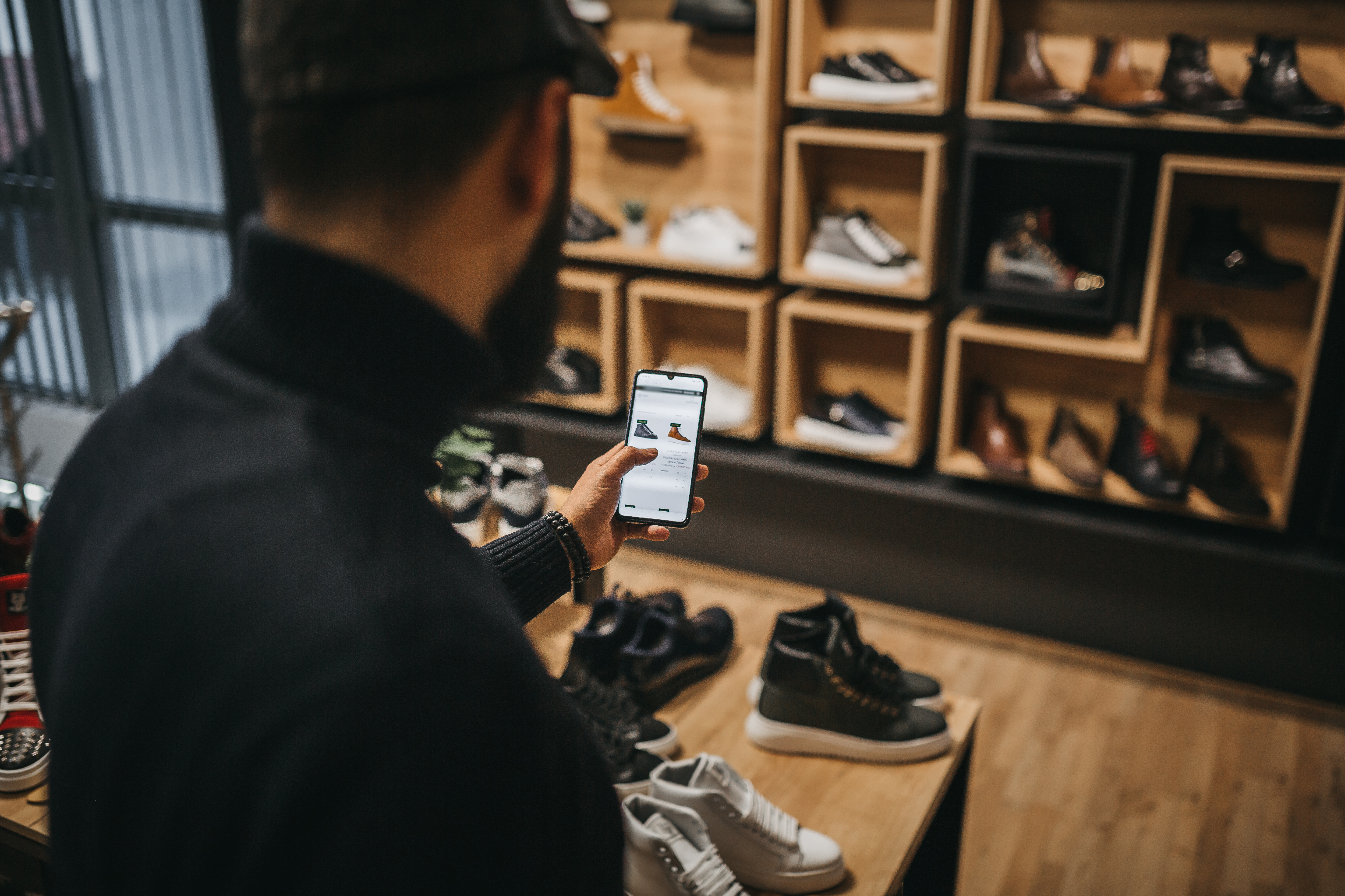 A man with a beard examines shoe options on his smartphone in a stylish shoe store, with various sneakers displayed in the background.